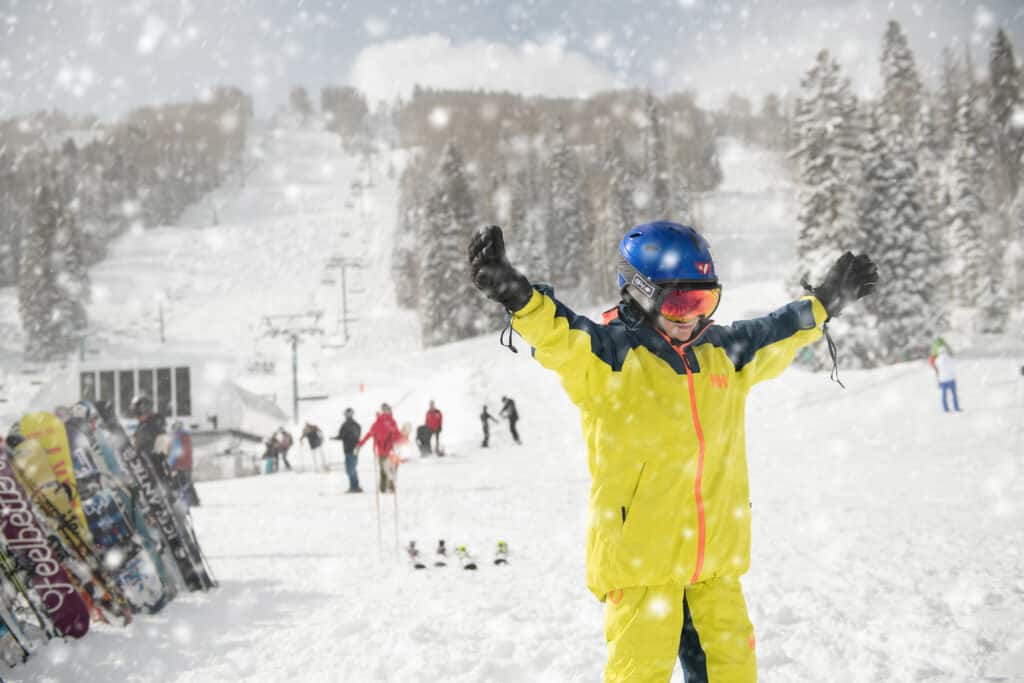 A kid in a yellow ski suit lifts his arms in the falling snow flakes