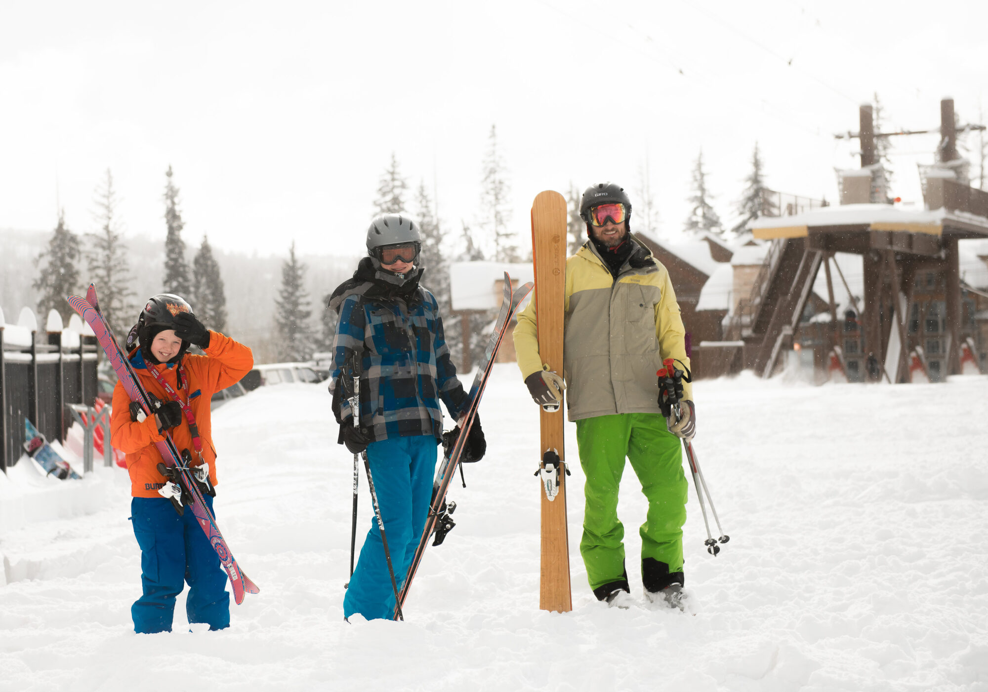 A family smiles with their skis in front of the ski lift at the base area