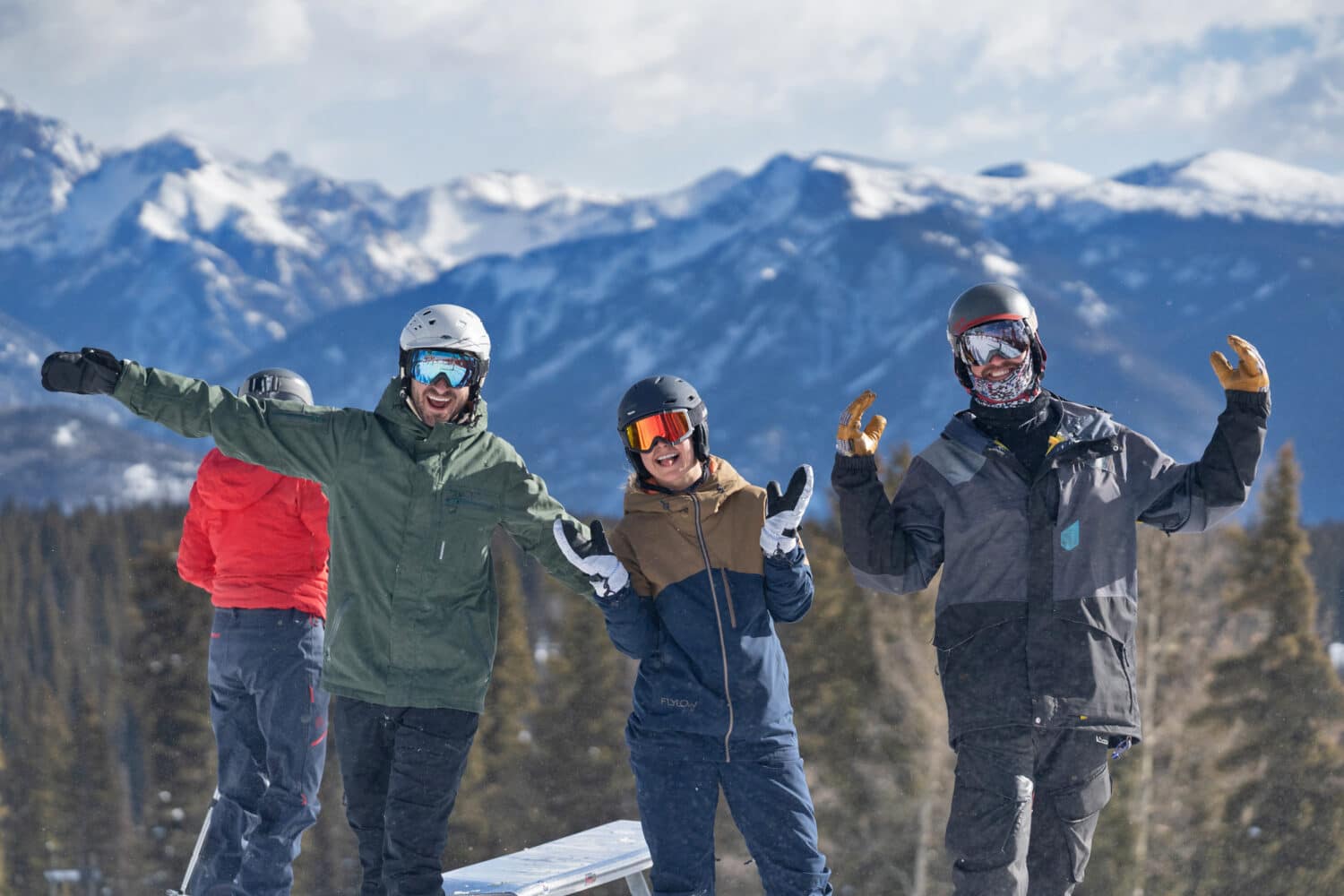Friends in ski gear smile with arms raised in front of a wintery mountain scene