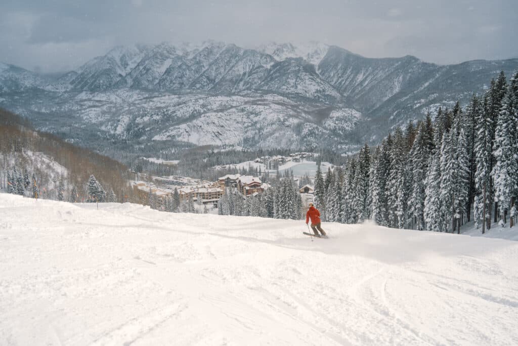 A skier in orange heads down Demon toward the base area with mountains in the background