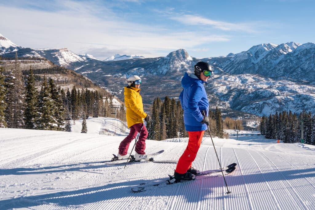 Two skiers smile at the top of a groomer run