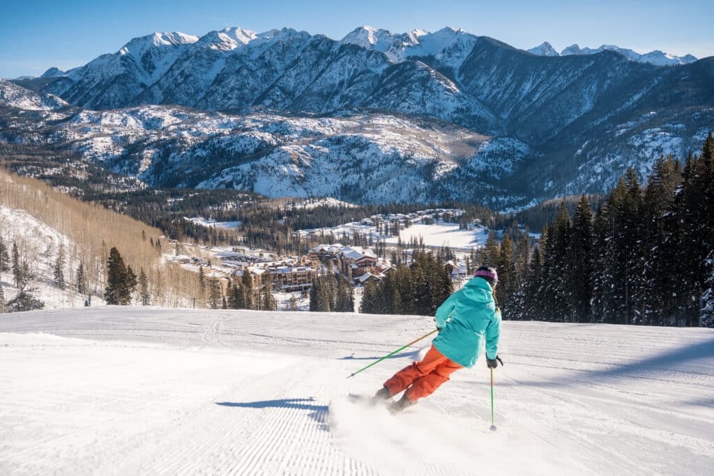 a skier in blue and orange skis on a groomer on a bluebird day toward mountains