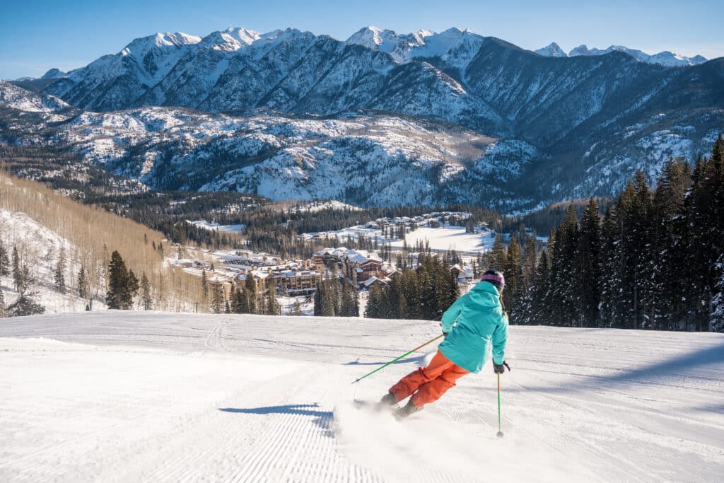 A skier in light blue jacket and orange pants skis a groomer with mountains in the background