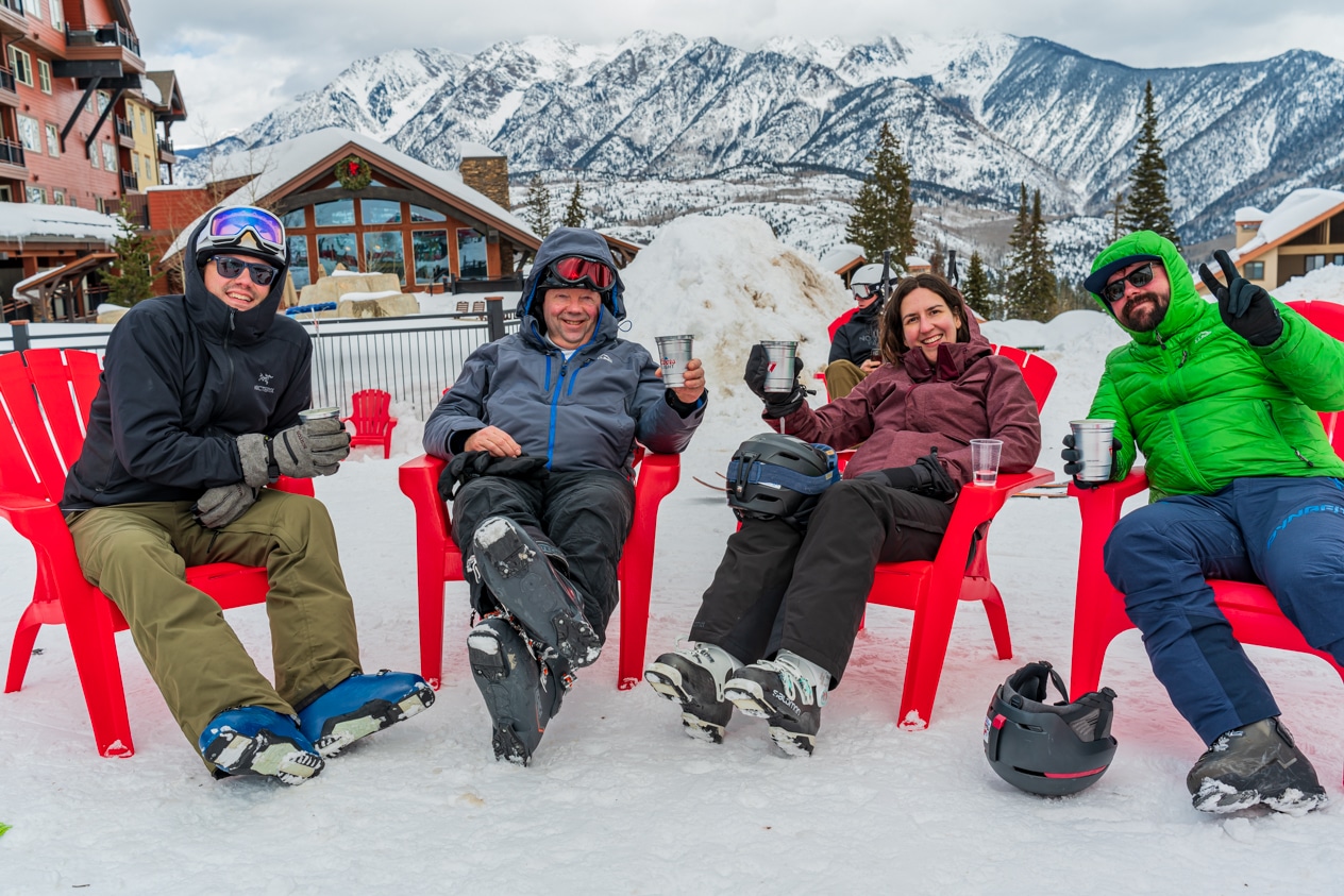 People in ski gear sit in red adirondack chairs with drinks on the ski beach at Purgatory Resort