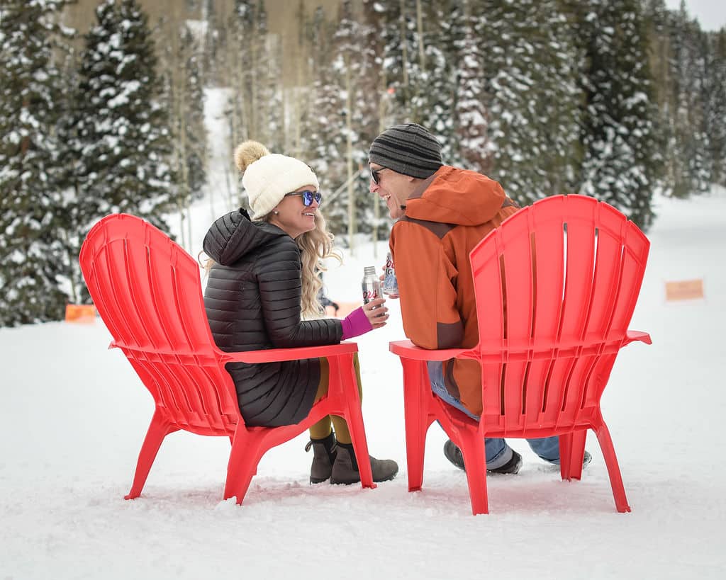 A man and woman smile at each other in red Adirondack chairs in the snow