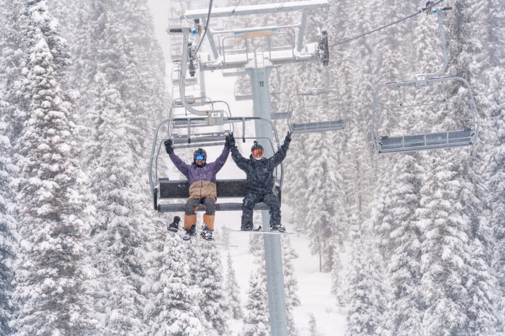 Two snowboarders raise their arms in celebration on the ski lift on a snowy day