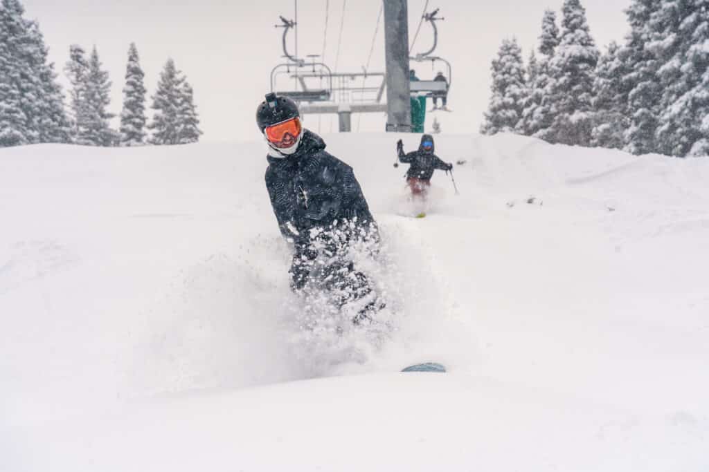 A snowboarder and skier cruise over fresh powder under a ski lift