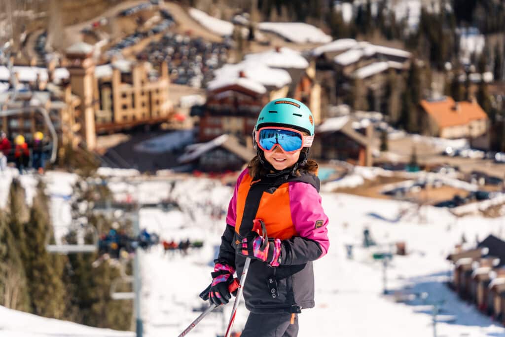 A child in a pink, orange and black ski jacket and turquoise helmet smiles from the slopes with the resort base area in the backgroune