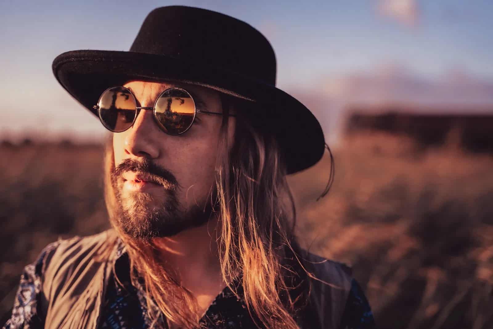 A portrait of Justin Larkin, a man with long brown hair and a trimmed beard with John Lennon glasses and a flat brim western style hat, outside at sunset in a desert setting.