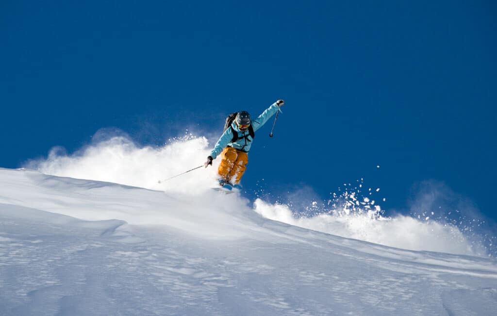 A skier in blue and orange gets some air on a powdery slope. Blue sky in the background. Photo Credit La Parva Ski Resort